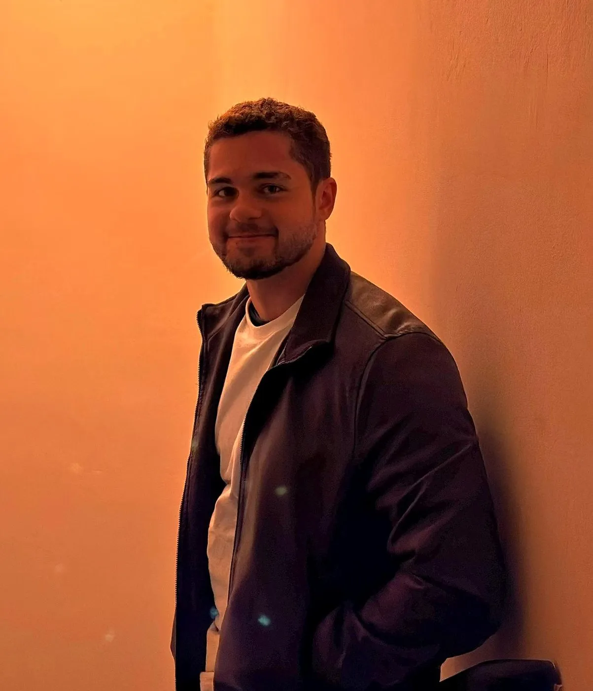 Enzo Gagliardi sorrindo, de jaqueta escura, numa parede iluminada por luz âmbar