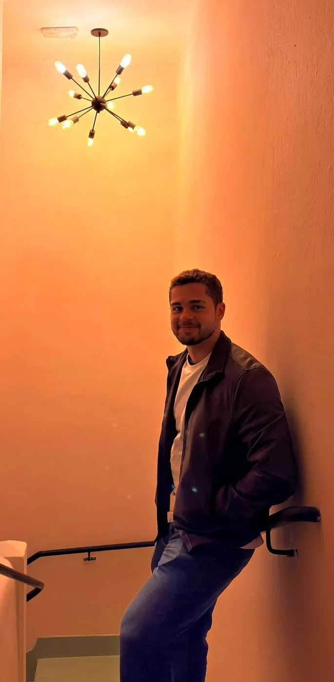 Enzo Gagliardi encostado na parede de uma escada iluminada por um lustre de luz âmbar